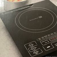 Top view of the Kuraidori induction hot plate in its original packaging showing the cooktop surface and control buttons.