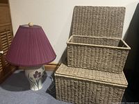 Photo showing a floral base table lamp with a purple pleated lampshade, next to two stacked wicker baskets with hinged lids.