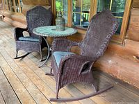 Two brown wicker rocking chairs with dark cushions and a round glass top table with metal legs and wicker trim on a wooden porch. A decorative lantern is on the table.