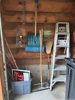 Photo showing snow shovels, brooms, a rake, and two metal step ladders against a wooden wall near garage area.
