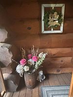 Wooden barrel planter filled with mixed artificial flowers in pink, white and dried beige tones, with small dog figurine beside it on the porch floor, rustic wood wall in background.