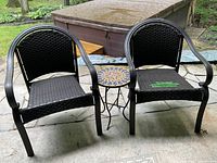 Two black woven patio chairs with metal frames positioned around a small round accent table with a colorful mosaic tile top on a stone patio.