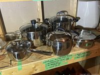Six stainless steel pots arranged on a wooden shelf, some with glass lids, showing a variety of sizes including large stock pots and smaller saucepans. Handles are metal or black plastic. Stickers with brand and size details visible on the shelf.