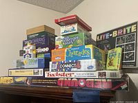 Large assortment of puzzles and board games stacked on a shelf including Lite-Brite, Family Charades, Connect 4, Twister, Super Termin8r, Dance Charades, and others.