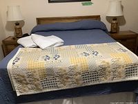 Full view of queen bed with mattress, blue fitted sheet, white linens and patterned quilt, flanked by two nightstands