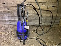 Full view of blue Simoniz S/400 pressure washer with coiled power cord, spray gun, and attached hose on a dirt floor in a barn environment.
