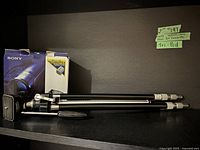 Photo showing Sony Handycam Mini DV Camcorder original box and a black and silver tripod laid on a shelf with handwritten identifying tags.