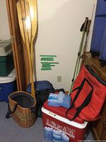 Wooden paddles, woven fishing basket, sleeping bags, walking poles, cooler with ice packs, and red insulated bag against wall with furniture