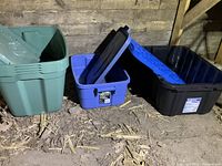 Three plastic storage bins with lids arranged on dirt floor inside a storage barn. One green, one blue, one black bin with lids visible.