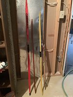 Three bows and assorted arrows standing against a wall in a basement, showing the colors and types of bows and arrows included.
