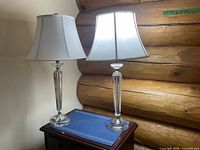 Two glass table lamps on a wooden table against a log cabin wall, showing overall condition and size.