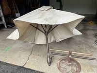 Full view of the beige outdoor sun umbrella next to the metal rolling stand, showing umbrella fabric and frame.