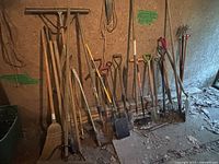 Wide shot showing 15 assorted gardening and yard tools such as brooms, rakes, shovels, hoes, and forks leaning against a barn wall on a dusty floor with cobwebs and debris.
