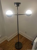 Full view of black metal floor lamp with two illuminated glass shades, standing in a corner against white walls and wood flooring.