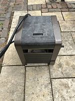 Photo showing the hose storage unit from top and side angle on stone pavement ground with visible wear on black top panel and brown metal side frame.