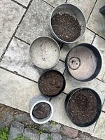 Six plastic planters arranged on stone pavement, showing top views with some soil inside, various sizes and colors gray and black.