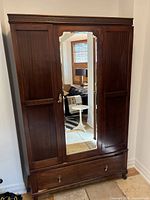 Front view of traditional dark brown wooden wardrobe with three doors, center door with full-length mirror and a bottom drawer with metal handles