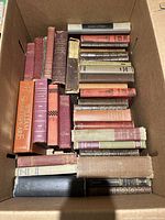Top-down view of assorted hardcover books with red, purple, orange, and beige covers tightly packed inside a box.