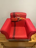 Red plush child's armchair featuring the Cars movie racing car graphic, suitable for play or rest.