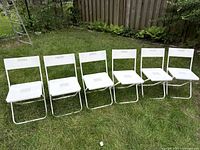 Six white folding chairs lined up on grass outdoors in a single row showing front view.