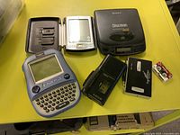Photo of assorted electronics including Palm Pilot, Sony Discman, iQuest organizer, two voice recorders, and VHS-C adapter cassette on yellow surface.