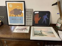 Wide view of the five wall hangings on wooden dresser surface including framed buffalo print, barbed wire panel, crosswalk sign, Indian Motorcycles sign and beach painting