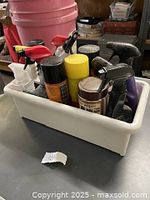 Overall view of assorted automotive cleaning and detailing supplies in a white plastic bin, showing spray bottles, aerosol cans, and other containers.