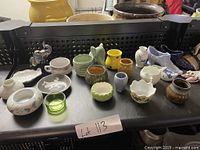 Photo showing multiple small decorative ceramic and glass items including bowls, plates, elephant figurines, and shoes spread out on a black surface.