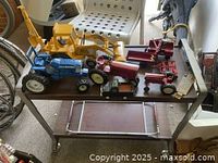 Overview shot of various die cast toy tractors and farm machinery lined up on table with ruler and chairs visible.
