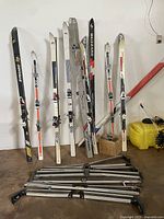 Multiple pairs of vintage downhill skis from brands including Dynamic, Rossignol, Blizzard, and Salomon leaning against a white wall, with roof racks laid out in front on the floor.