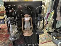 Three Dietz oil lamps hanging on black pegboard: two black in good condition, one rust-colored in middle with missing cap