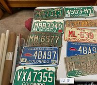 Photo showing assorted vintage Colorado license plates arranged on a table, including green and blue plates and a yellow plate partially visible
