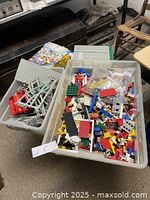 Two plastic bins filled with assorted vintage LEGO bricks, train tracks, and base plates. Visible minifigures and instruction booklets beside the bins.