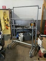 Pair of galvanized pipe double-bar clothing racks with rolling castors positioned indoors among various other items.
