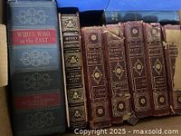 Photo of antique books showing Who's Who in the East by A.N. Marquis, Henry Wadsworth Longfellow book, and multiple volumes of George Eliot books in brown leather with gold embossing.