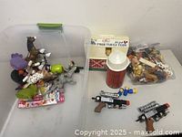 Photo showing a clear plastic bin filled with various toy animals and figurines, a small red-white barn structure, a red cylindrical silo, and a plastic bag with additional figures and two black and silver foam dart blasters.