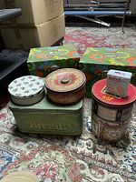 Photo showing the full group of vintage cookie and storage tins arranged on a patterned rug with two rectangular floral print tins in the background.
