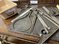 Top-down view of vintage machinist and drafting tools arranged on a work surface, shows calipers, dividers, combination squares, rulers, and drafting arm.
