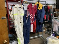 Five pieces of vintage outerwear on hangers including ski suits, jackets, and motorcycle jacket displayed on a metal rack.