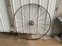 Full view of an antique wagon wheel with a metal rim and multiple thin metal spokes. The wheel measures approximately 54 inches in diameter. Some spokes are bent and broken, rim shows rust and wear.