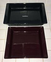 Two ceramic baking pans placed side by side, black rectangular and burgundy square, both glossy finish with Emile Henry branding on the black pan.