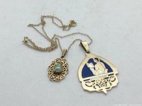 Two gold-filled pendants on a gold tone chain, one with an opal stone in ornate filigree and the other with a blue enamel figure.