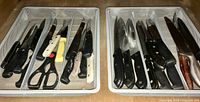 Two white wire cutlery trays containing various knives and a pair of kitchen scissors divided into compartments.