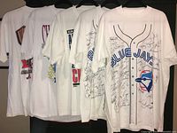 Five vintage white Toronto Blue Jays T-shirts hanging, showing front designs and multiple autographs in black marker covering the front of each shirt.