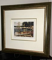 Framed print in dark wood and gold detailed frame shown angled, depicting a canoe on a lake shore