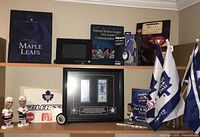 Wide view of two shelves containing multiple Toronto Maple Leafs hockey collectibles including books, framed memorabilia, bobbleheads, flags, and paper signs.