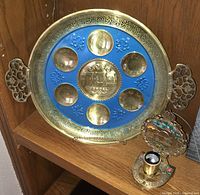 Brass Judaica Seder plate with seven sections and Hebrew inscriptions, bright blue enamel background, set on a wooden shelf next to a small decorative table lamp with a floral design.