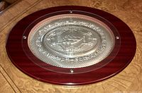 Oval serving tray with embossed sterling silver center mounted on a reddish-brown wooden base.