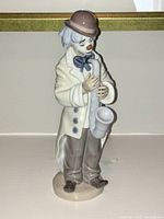 Front view of Lladro porcelain clown figurine playing saxophone, showing detailed paint including brown hat, blue bow tie, cream coat, and brown pants.