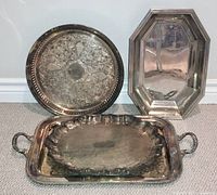 Four vintage/antique silver plated trays displayed together on the floor showing varied shapes and design details including engraving on one tray